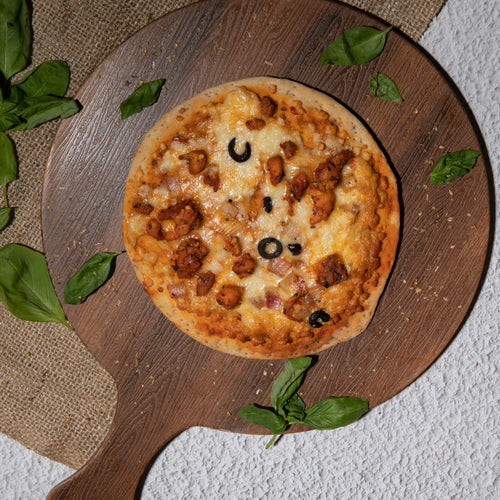 Chicken pizza kept upon a wooden pizza platter with some basil leaves aside kept upon a jute mat.