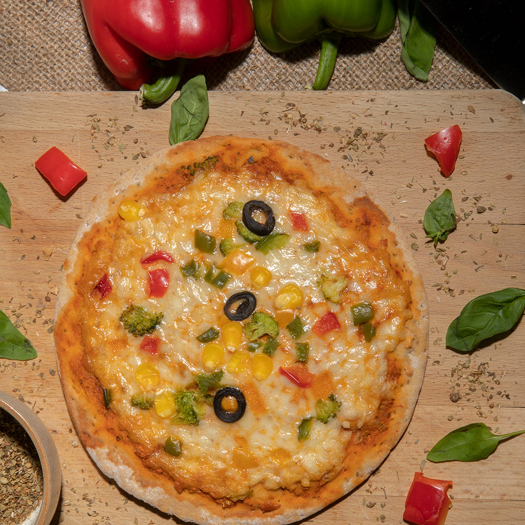 A delicious pizza kept upon a wooden platter with some basil leaves, oregano and bell peppers aside kept upon a jute mat.